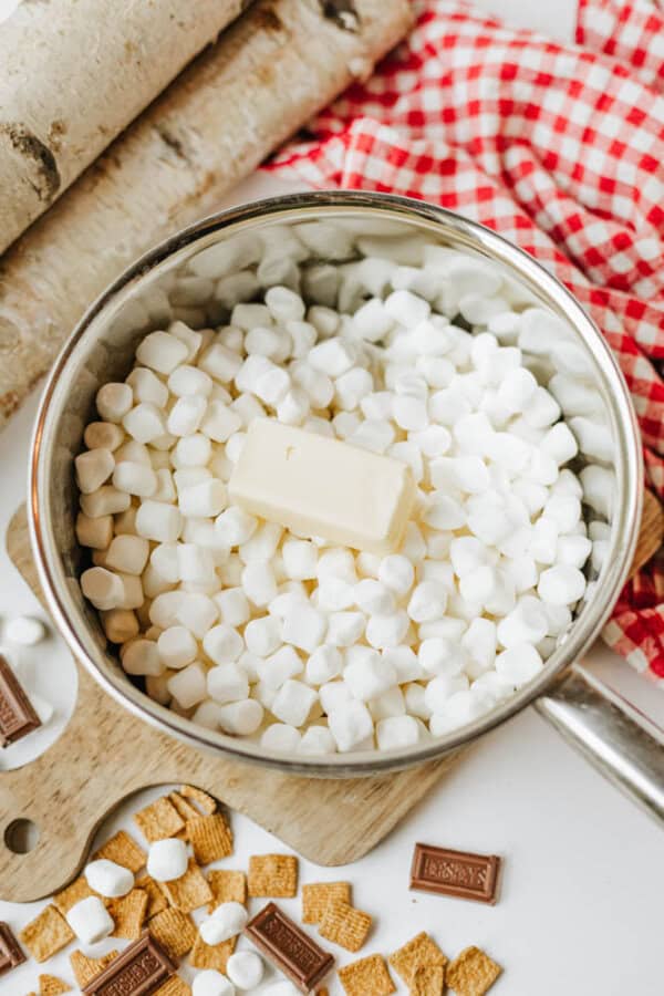A saucepan filled with mini marshmallows and a stick of butter sits on a white surface beside a red checkered cloth, chocolate pieces, graham cereal, and birch logs.