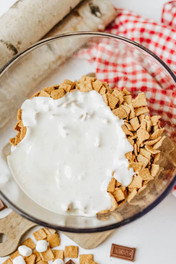 A glass bowl filled with cinnamon cereal squares topped with melted marshmallow mixture. Pieces of chocolate, mini marshmallows, and a red checkered cloth are nearby on a white surface.