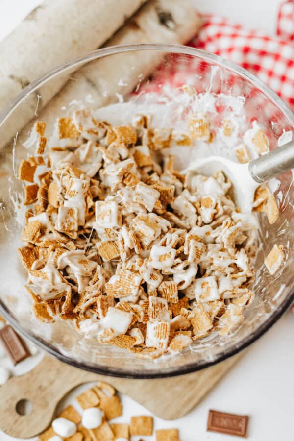 A glass bowl filled with Chex cereal mixed with melted marshmallow and a white spatula, surrounded by graham crackers, marshmallows, and chocolate on a wooden board.