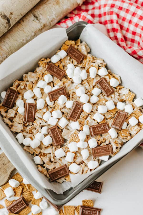 A square baking pan lined with parchment paper is filled with smores bars made of mini marshmallows, graham cereal, and pieces of Hersheys chocolate, with extra marshmallows and chocolate scattered on top.