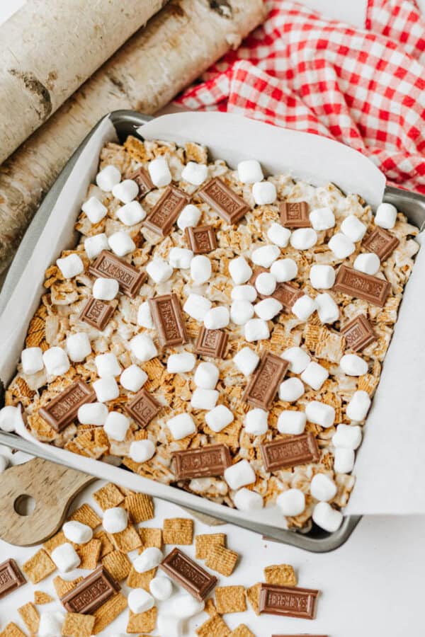 A square baking pan filled with smores bars topped with mini marshmallows and pieces of Hersheys chocolate, sitting on parchment paper. Nearby are graham cereal pieces and more chocolate on a white surface.
