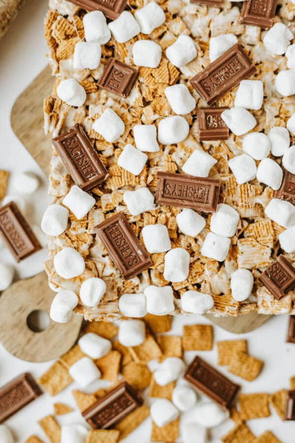 A close-up of dessert bars topped with mini marshmallows, square pieces of Hershey’s chocolate, and golden cereal pieces on a wooden board, with extra toppings scattered around.
