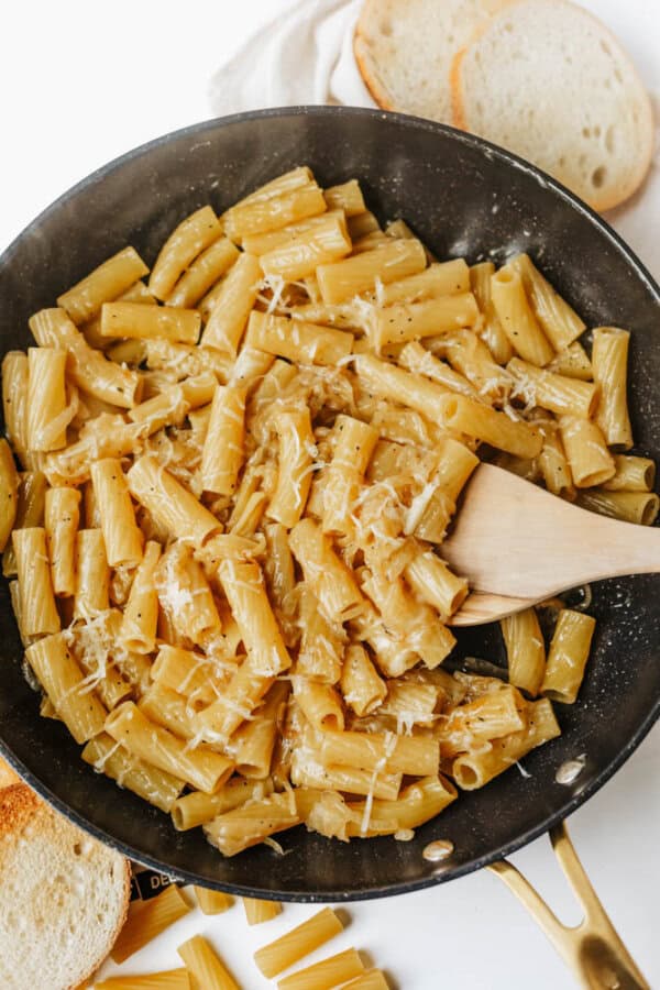 A black pan filled with cooked rigatoni pasta coated in a light sauce, topped with grated cheese. A wooden spatula stirs the pasta, and slices of bread are nearby on a white surface.