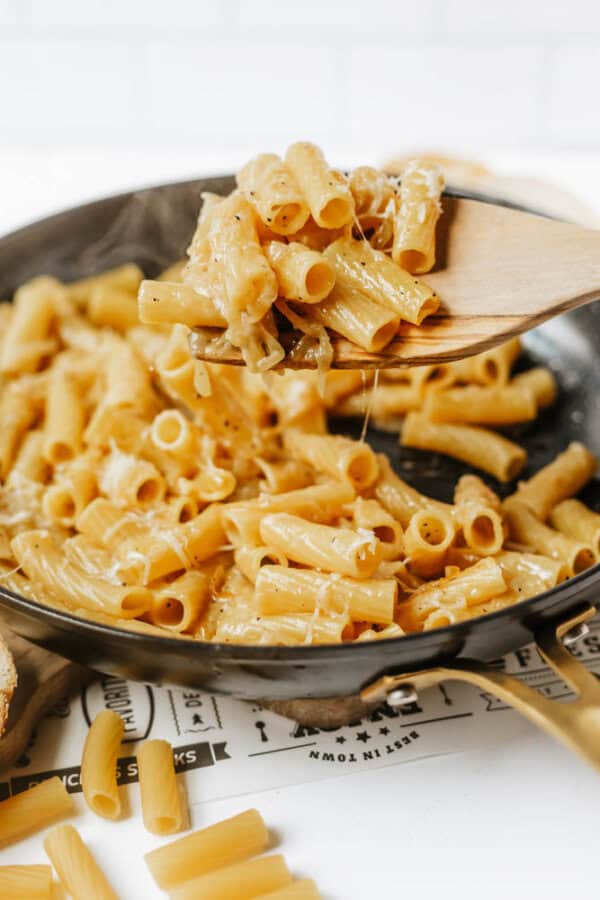 A wooden spoon lifts cheesy rigatoni pasta from a skillet, with melted cheese stretching between the spoon and the pasta. Uncooked rigatoni pieces are scattered on the table below.