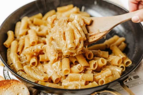 A close-up of creamy rigatoni pasta being stirred with a wooden spoon in a black skillet, with melted cheese coating the pasta. A slice of toasted bread is visible in the corner.