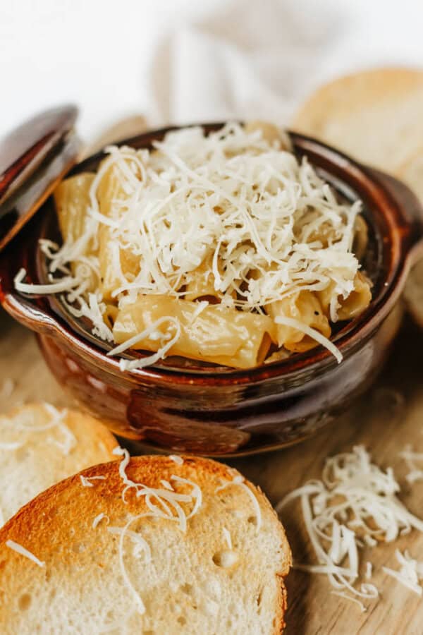 A brown ceramic bowl filled with creamy macaroni and cheese, topped with shredded cheese, is surrounded by slices of toasted bread on a wooden surface.