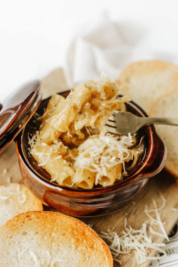 A brown ceramic bowl filled with cheesy pasta, topped with shredded cheese, sits on a wooden board next to slices of toasted bread. A fork lifts some pasta from the bowl.