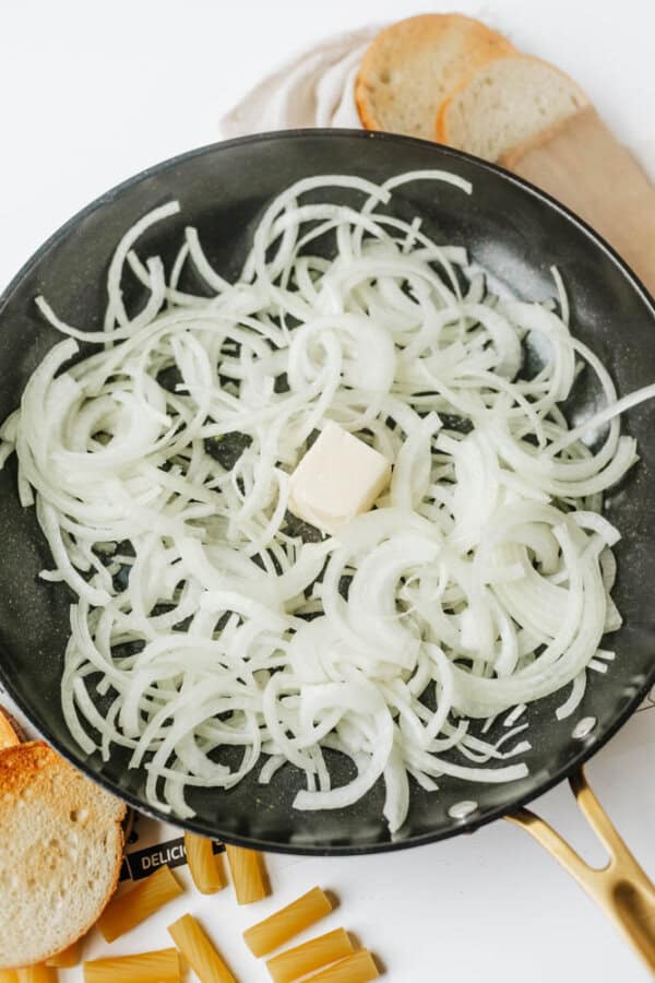 A skillet with sliced onions and a pat of butter in the center, ready to be cooked. Toasted bread slices and uncooked pasta are visible around the pan.