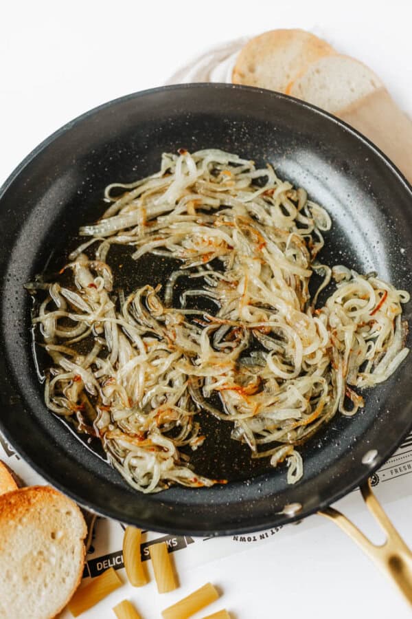 A black skillet with thinly sliced, caramelized onions sits on a white surface. Toasted bread slices and some uncooked pasta are visible around the pan.