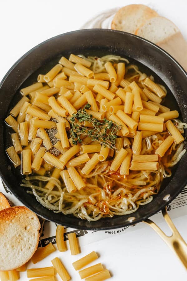 A skillet with uncooked rigatoni pasta, sprigs of thyme, seasonings, and broth over sautéed onions. Slices of toasted bread and scattered pasta are nearby.