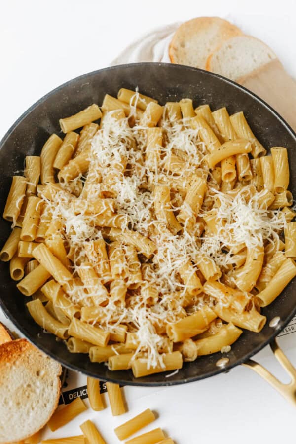 A skillet filled with cooked rigatoni pasta topped with grated cheese, with slices of toasted bread and uncooked pasta pieces nearby on a white surface.