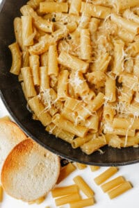 A close-up of a black pan filled with rigatoni pasta coated in a creamy sauce and sprinkled with grated cheese. Two slices of toasted bread and a few uncooked pasta pieces are on the white surface nearby.