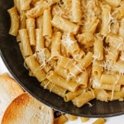 A close-up of a black pan filled with rigatoni pasta coated in a creamy sauce and sprinkled with grated cheese. Two slices of toasted bread and a few uncooked pasta pieces are on the white surface nearby.