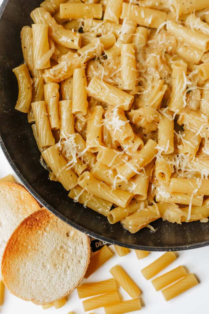 A close-up of a black pan filled with rigatoni pasta coated in a creamy sauce and sprinkled with grated cheese. Two slices of toasted bread and a few uncooked pasta pieces are on the white surface nearby.