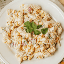 A plate of creamy street corn salad with pasta, corn, red onion, and cilantro sits on a white plate, garnished with fresh cilantro. The text reads “Street Corn Salad Recipe” and “homemadeheather.com.”.