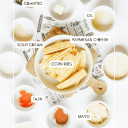 A flat lay of labeled ingredients for corn ribs, including corn, cilantro, butter, oil, parmesan cheese, sour cream, mayo, tajin, seasoning, and sugar in small bowls on a white background.