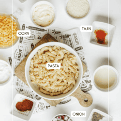 A flat lay of ingredients in bowls on a white surface, including pasta, corn, sour cream, Tajin seasoning, chopped onion, spices, and herbs, each labeled with text.