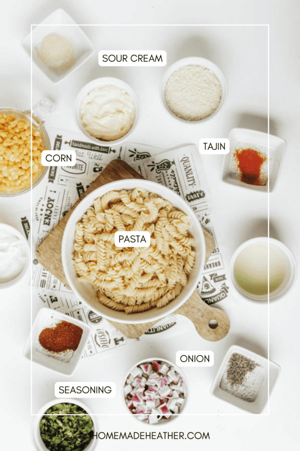 A flat lay of ingredients in bowls on a white surface, including pasta, corn, sour cream, Tajin seasoning, chopped onion, spices, and herbs, each labeled with text.