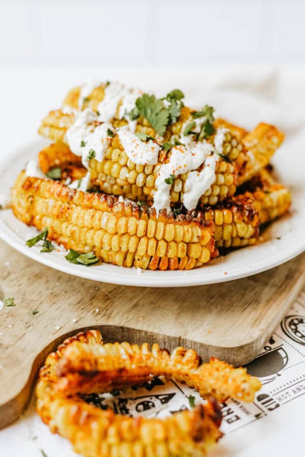 A plate of grilled corn ribs topped with creamy sauce and chopped cilantro, served on a wooden board with extra corn ribs beside it.
