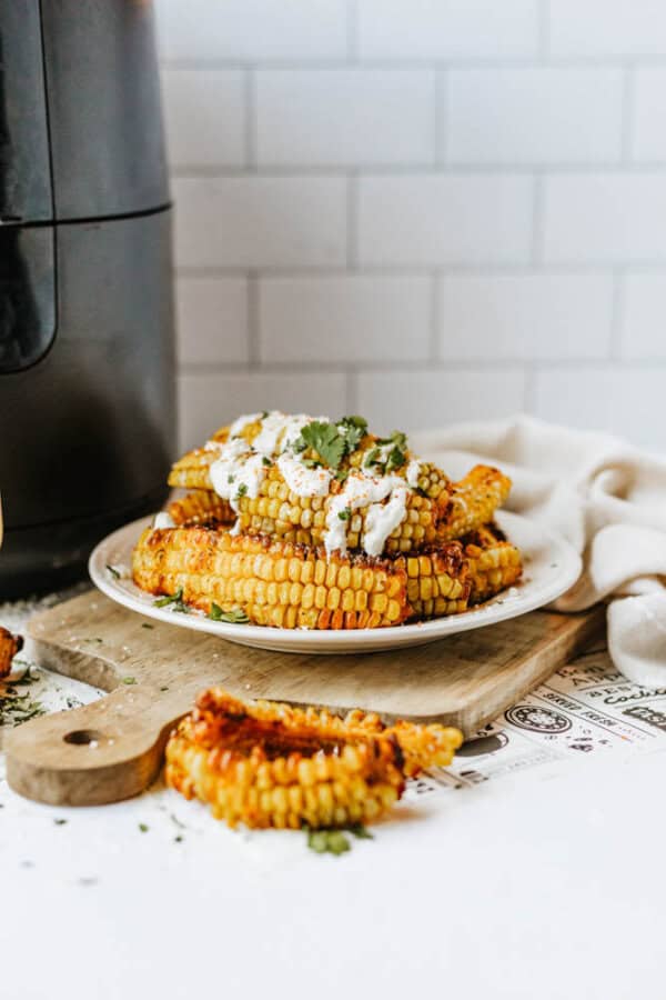 A plate of grilled corn ribs topped with a creamy sauce and fresh herbs sits on a wooden board, with an air fryer and white tiled wall in the background. A piece of corn rib rests in the foreground.