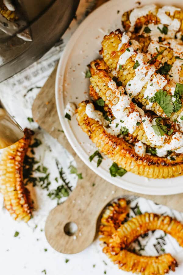 A plate of roasted corn ribs drizzled with a creamy white sauce, sprinkled with chopped cilantro and seasoning, sits on a wooden cutting board with additional corn ribs nearby.