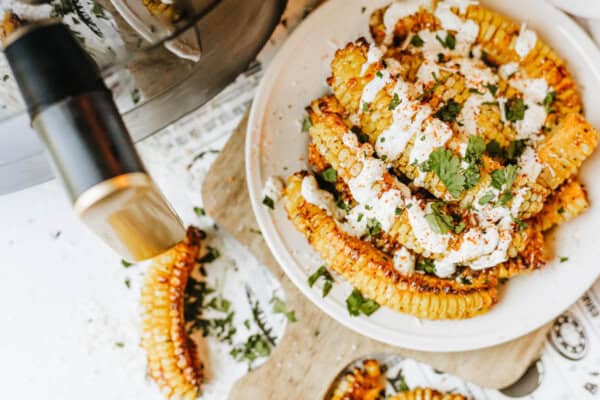 A plate of grilled corn ribs topped with creamy sauce, chopped cilantro, and spices, with some corn and sauce spilled on a white surface nearby. A kitchen appliance is partially visible on the left.