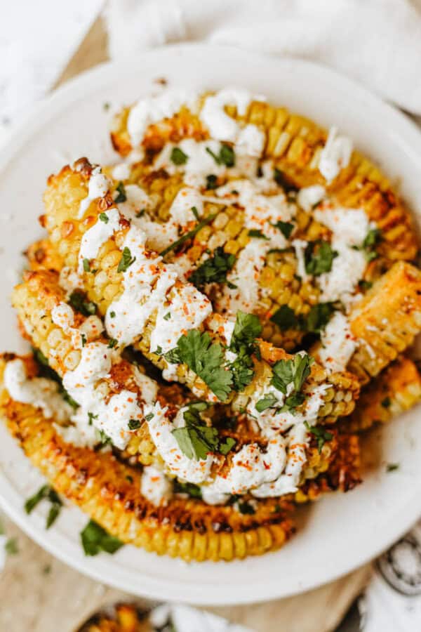 A plate of grilled corn ribs topped with a creamy white sauce, chopped cilantro, and a sprinkle of red seasoning. The corn is cut into rib-like strips and arranged on a white plate.