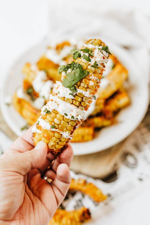 A hand holds a piece of grilled corn rib drizzled with creamy sauce and topped with cilantro, with more corn ribs on a plate in the background.