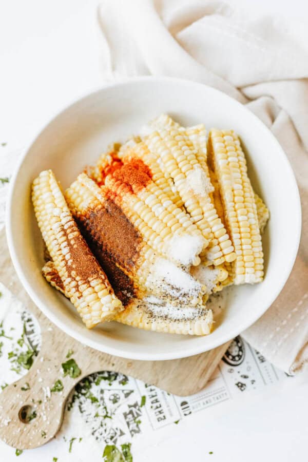 A white bowl filled with uncooked corn on the cob pieces, sprinkled with various spices including paprika, chili powder, salt, and pepper, sits on a wooden board beside a light-colored cloth.