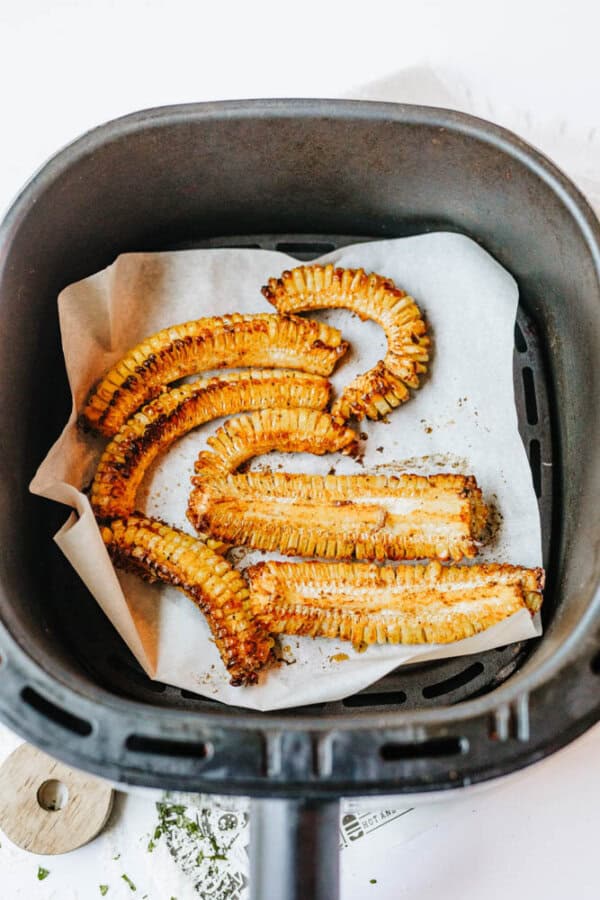Curly, golden-brown air-fried enoki mushrooms on parchment paper inside an air fryer basket, with slightly crispy edges and seasoning visible.