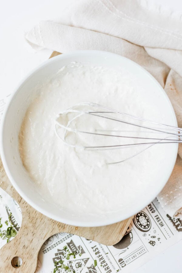 A white bowl filled with thick batter being mixed with a metal whisk, placed on a wooden cutting board next to a beige cloth.