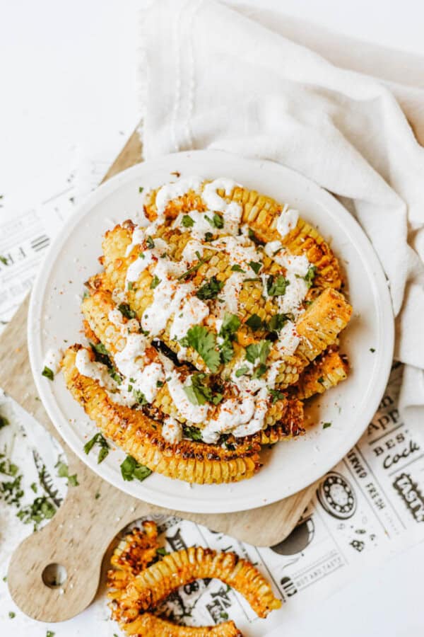A plate of grilled corn ribs garnished with creamy white sauce, chopped cilantro, and spices, served on a white dish atop a wooden board and cloth napkin, with extra cilantro sprinkled around.