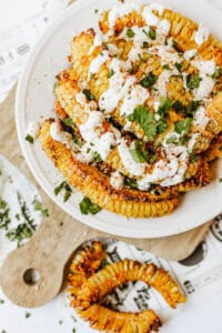 A plate of roasted corn ribs topped with a creamy white sauce, chopped cilantro, and spices, served on a cutting board with extra herbs scattered around.