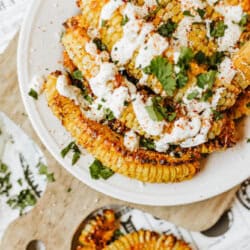 A plate of roasted corn ribs topped with a creamy white sauce, chopped cilantro, and spices, served on a cutting board with extra herbs scattered around.
