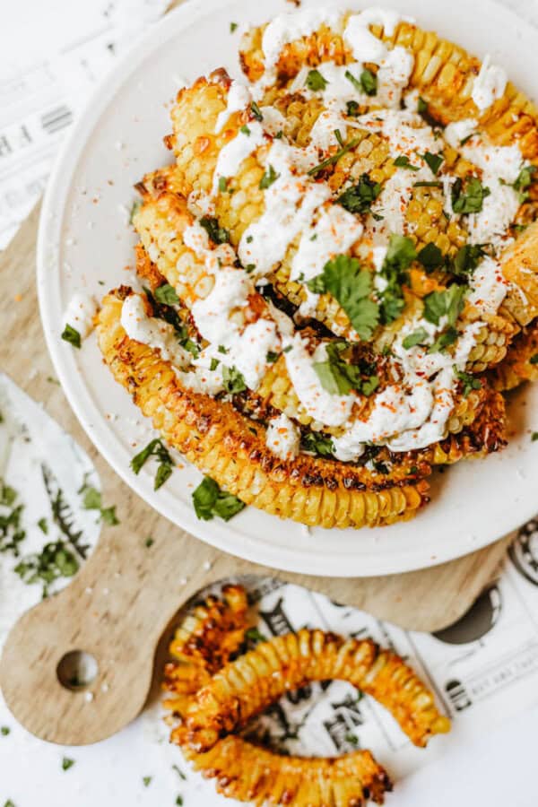 A plate of roasted corn ribs topped with a creamy white sauce, chopped cilantro, and spices, served on a cutting board with extra herbs scattered around.
