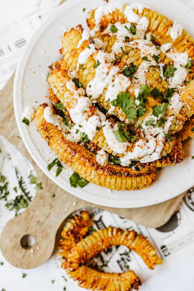 A plate of roasted corn ribs topped with a creamy white sauce, chopped cilantro, and spices, served on a cutting board with extra herbs scattered around.