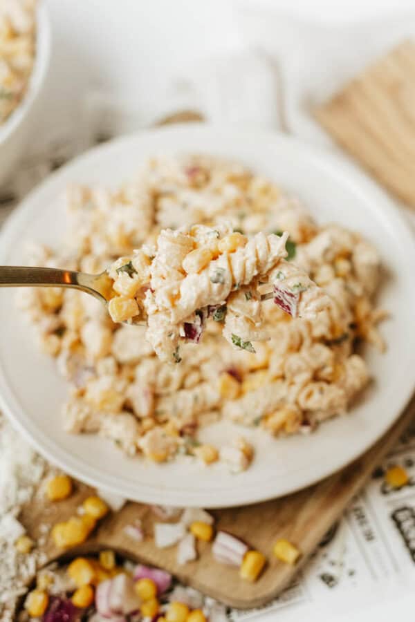 A close-up of a fork holding a bite of creamy pasta salad with corn, red onion, and herbs above a plate filled with the same salad.