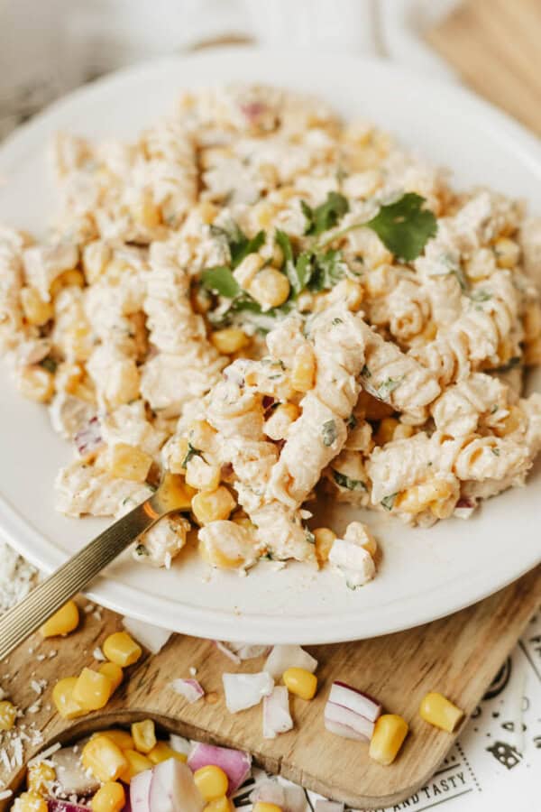 A plate of creamy pasta salad with spiral noodles, corn, chopped red onion, and fresh herbs, garnished with a sprig of cilantro. A fork rests on the side, with some ingredients scattered on the wooden surface nearby.