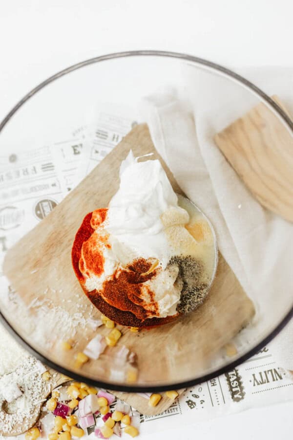 A glass bowl containing sour cream, mayonnaise, and various spices, sits on a wooden board next to a towel and scattered ingredients including flour, corn, and diced onions.