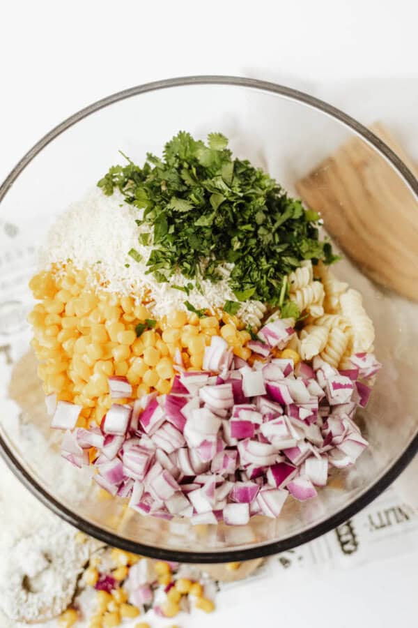 A glass bowl filled with uncooked pasta, chopped red onion, corn, shredded cheese, and fresh chopped cilantro, arranged in separate sections before mixing.
