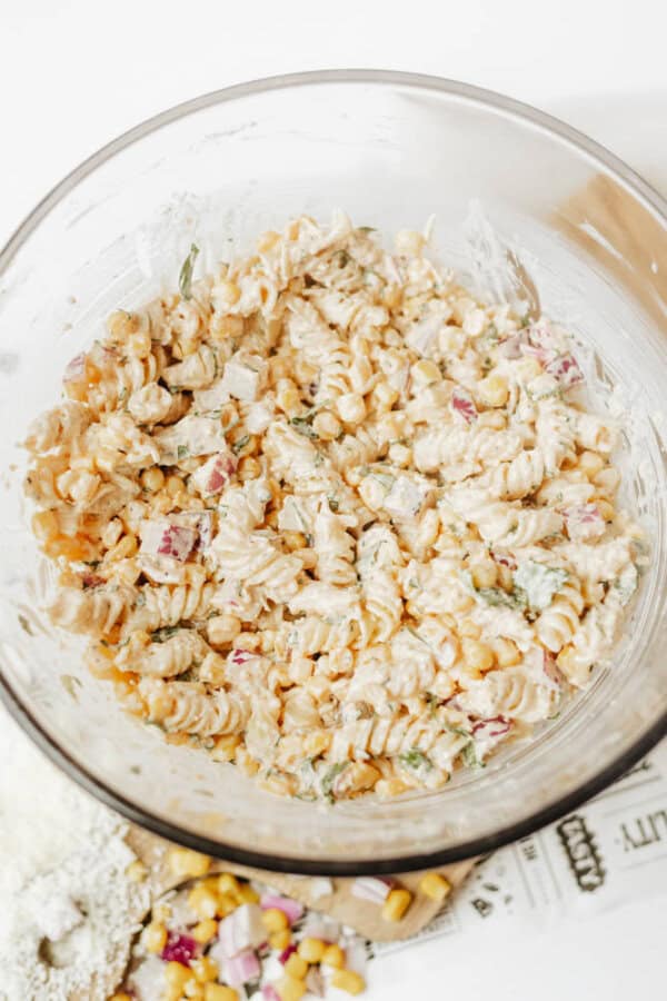 A glass bowl filled with creamy pasta salad made of rotini pasta, corn, chopped red onion, and herbs. Ingredients like corn and grated cheese are scattered on a surface nearby.