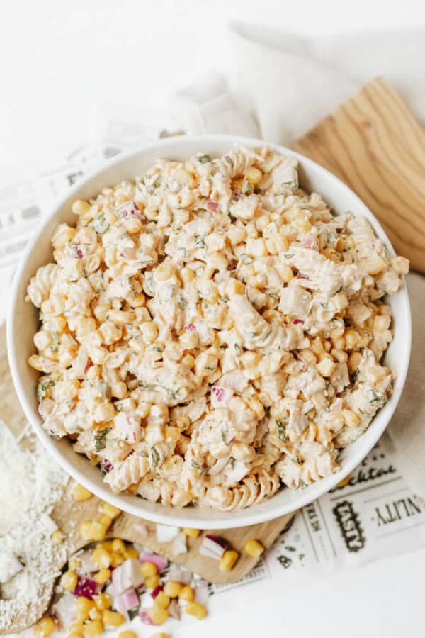 A white bowl filled with creamy pasta salad containing rotini, corn, chopped red onion, and herbs, placed on a table with some spilled corn and onions nearby and newspaper underneath.