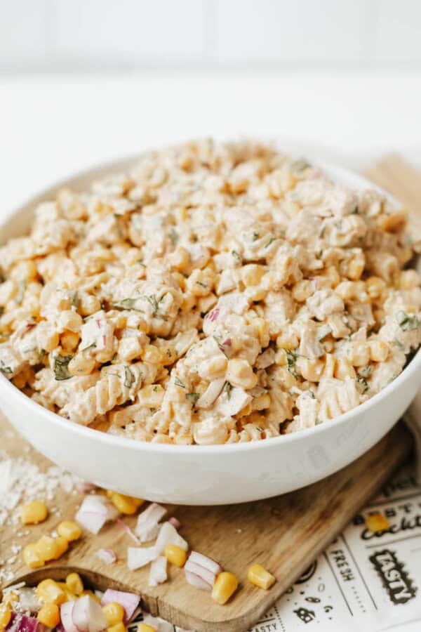 A white bowl filled with creamy pasta salad featuring rotini noodles, corn, red onion, and fresh herbs, sitting on a wooden board with scattered corn kernels and chopped onions nearby.