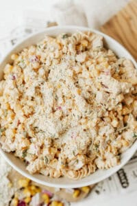 A bowl filled with creamy pasta salad made of rotini pasta, corn, diced red onion, herbs, and topped with grated cheese, sitting on a white surface.
