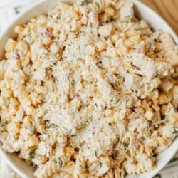 A bowl filled with creamy pasta salad made of rotini pasta, corn, diced red onion, herbs, and topped with grated cheese, sitting on a white surface.