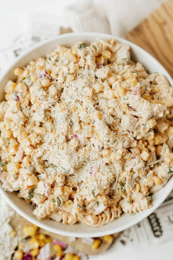 A bowl filled with creamy pasta salad made of rotini pasta, corn, diced red onion, herbs, and topped with grated cheese, sitting on a white surface.