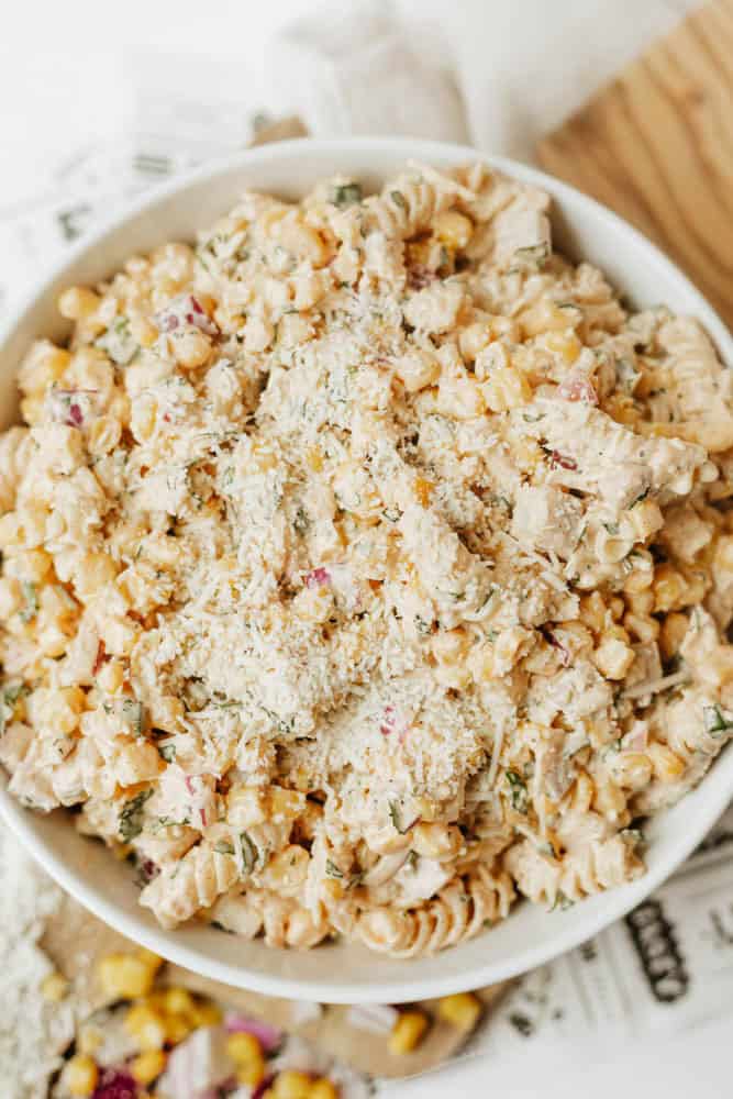 A bowl filled with creamy pasta salad made of rotini pasta, corn, diced red onion, herbs, and topped with grated cheese, sitting on a white surface.