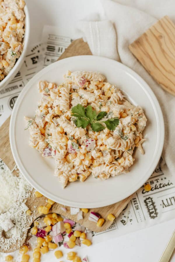 A white plate with a serving of creamy pasta salad containing corn, red onion, and herbs, garnished with fresh cilantro, sits on a table set with a bowl of salad, corn kernels, and parmesan cheese.