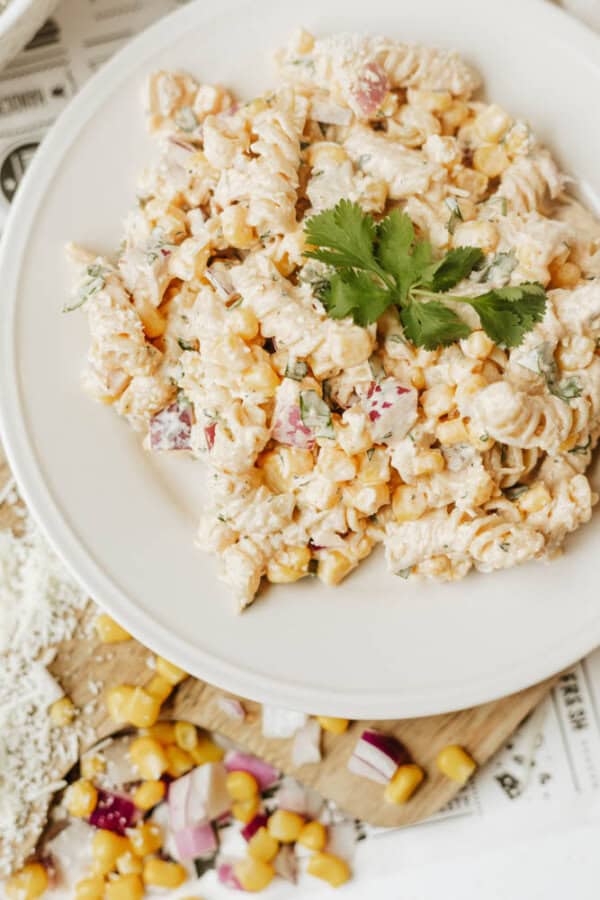 A white plate with a creamy pasta salad containing fusilli pasta, corn, diced red onions, cilantro, and cheese, garnished with cilantro. Some salad ingredients and cheese are scattered on a wooden board nearby.