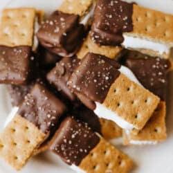 A plate of 3-ingredient S'mores Bites: rectangular graham cracker sandwiches filled with white marshmallow, partially dipped in chocolate, and sprinkled with coarse sea salt.